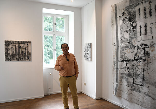 Till Richte steht in einem weißen Raum. Links und rechts hängen große Bilder an der Wand. Er lacht.