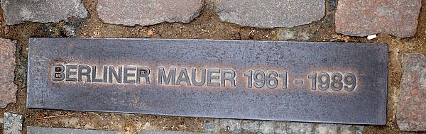 Auf Kopfsteinpflaster liegt ein Metallschild. Darauf steht "Berliner Mauer 1961-1989"