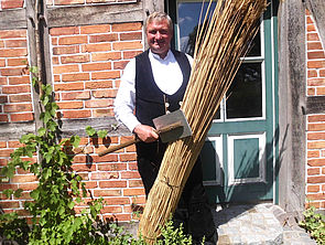 Reetdachdecker Joachim Schröter steht in schwarzer Arbeitshose, Weste und weißem Hemd vor einem Fachwerkhaus. In einer Hand hält er ein Bündel Reet. In der anderen ein Werkzeug. 