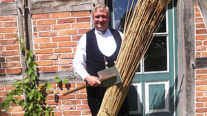 Reetdachdecker Joachim Schröter steht in schwarzer Arbeitshose, Weste und weißem Hemd vor einem Fachwerkhaus. In einer Hand hält er ein Bündel Reet. In der anderen ein Werkzeug. 