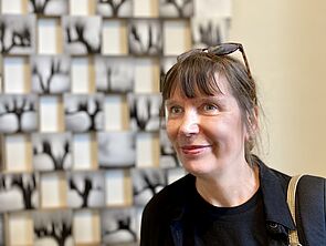 Petra Schröck steht vor einem Kunstwerk, das an der Wand hängt.