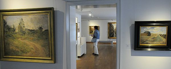 Links und rechts von einer offenen Tür hängt ein Bild. Im Raum dahinter steht eine Frau und schaut sich Gemälde an.