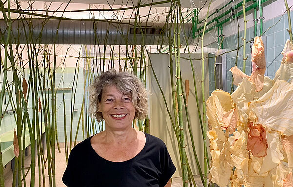 In einem Waschraum verteilen sich vom Boden bis zur Decke unzählige grüne, bambusartige Triebe. Dazwischen hängt eine Wagenrad große helle Blüte. Die Künstlerin steht mitten in ihrer Installation.