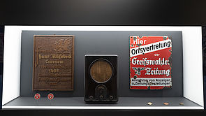 Eine Vitrine mit zwei Schildern und einem Radio aus der Vorkriegszeit des 2. Weltkrieges.