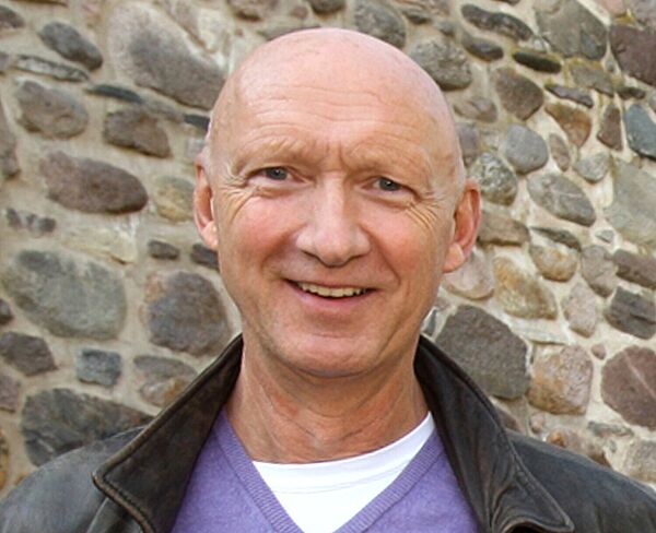 Porträt eines Mannes. Er blickt in die Kamera, trägt eine Jacke und einen fliederfarbenen Pullover.