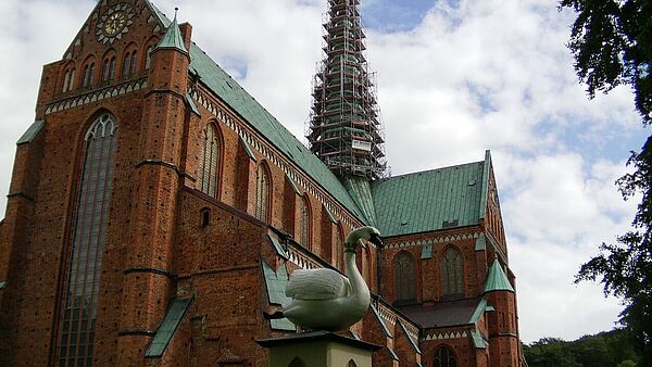 Ein Backsteingebäude in L-Form mit grünem Dach. Der Turm ist eingerüstet. Auf einem Sockel vor dem Gebäude steht ein Schwan.