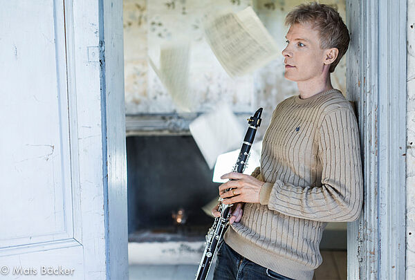 Ein junger Mann lehnt an einem Türrahmen. Er hält eine Klarinette in den Händen. Im Hintergrund fliegen Notenblätter durch die Luft.