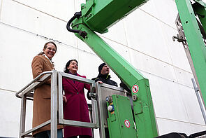 Zwei Frauen und ein Mann auf einem Hubsteiger. Sie fahren nach oben.