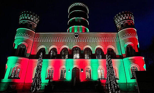 Ein Schloss. Das Schloss ist angestrahlt in Rot und Grün. Vor dem Schloss stehen zwei Weihnachtsbäume. Es ist dunkel.