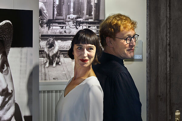 Katharins John steht Rücken an Rücken mit Manfred W. Jürgens. Im Hintergrund hängen Fotografien.