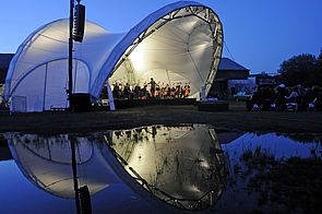 Eine beleuchtetes, muschelartiges Konzertzelt. Darin sitzt die Norddeutsche Philharmonie und spielt.