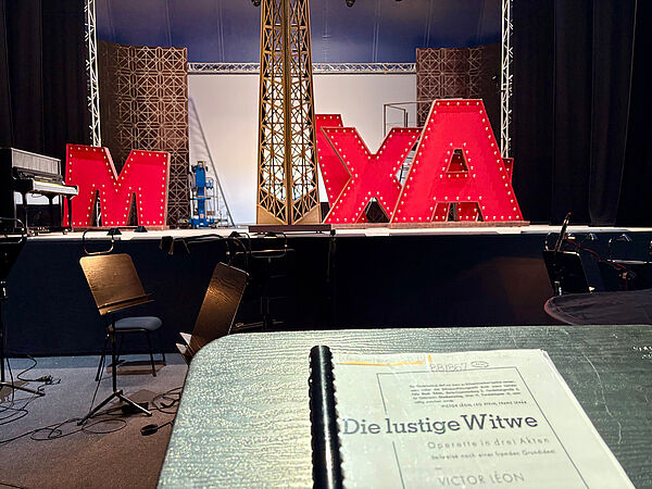 Ein Pult. Auf dem Pult liegt ein Heft. Auf dem Heft steht: "Die lustige Witwe". Dahinter ist eine Bühne. 