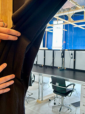 Ein Hand schiebt einen Vorhang beiseite. Dahinter ist ein Raum. In dem Raum stehen Tische und Stühle.