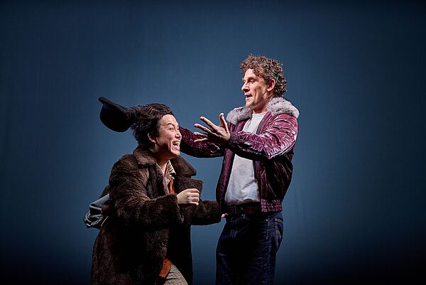 Ein Mann setzt einem anderen Mann einen Hut auf. Beide tragen Kostüme.