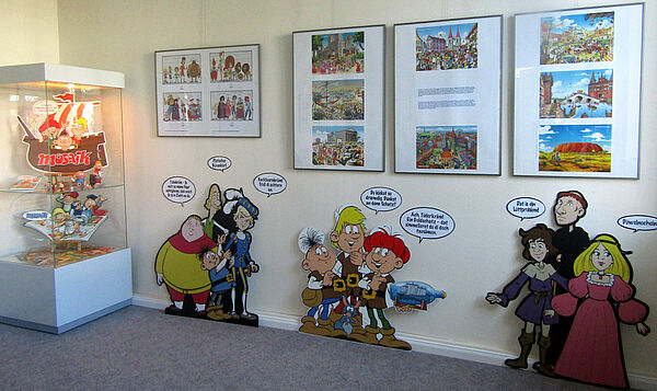 An einer Wand stehen Pappfiguren aus den Geschichten der Abrafaxe. Über ihnen hängen Bilderrahmen mit Szenen aus den Heften. Links steht eine Vitrine mit Mosaik-Exponaten.