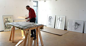 Der Künstler steht an einem Tisch und sortiert Zeichnungen. An den Wänden lehnen Bilderrahmen mit Kunstwerken.