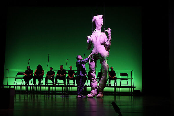Eine Bühne. Ein Mann steht für einer übergroßen Figur aus Stoff. Im Hintergrund sitzen Schauspieler nebeneinander.