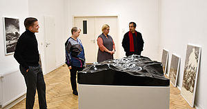 Auf einem weißen Sockel liegt eine schwarze Skulptur. Der Künstler und die Kunstkommission betrachten sie.