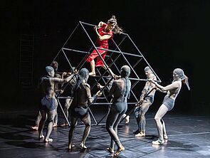 Eine Frau im roten Kleid klettert auf ein Gerüst, das von mehreren Leuten mit Strumpfmasken getragen wird.