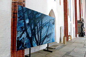 Ein großes Bild in blauen Farben steht vor einer Kirchenwand.