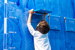 Ein Kind steht vor einer blauen Wand und schaut unter eine Textklappe. 