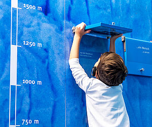 Ein Kind steht vor einer blauen Wand und schaut unter eine Textklappe. 