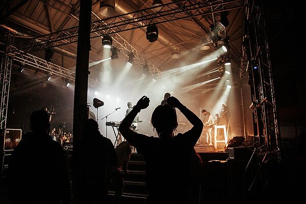 Eine Bühne. Auf der Bühne sind Musiker. Die Bühne ist beleuchtet. Vor der Bühne stehen Zuschauer. 