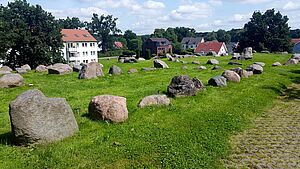 Auf einer Wiese liegen mehrere Findlinge. Im Hintergrund stehen Wohnhäuser.