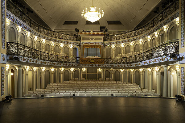 Ein Saal im Theater. Im Saal ist eine Bühne. Vor der Bühne stehen viele Stühle.