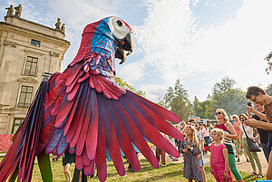 Eine Wiese. Auf der Wiese steht eine große Figur. Die Figur ist ein bunter Vogel und größer als Menschen. Neben der Figur stehen Menschen.
