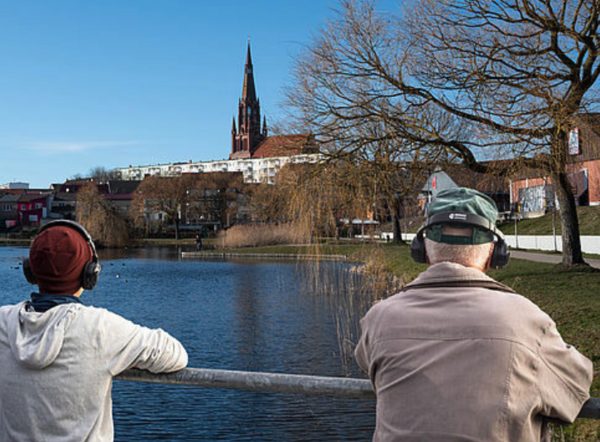 Ein Mann und ein Junge stehen an einem Geländer und blicken auf Demmin. Vor ihnen liegt Wasser, am Ufer stehen Häuser. Die beiden tragen Kopfhörer.