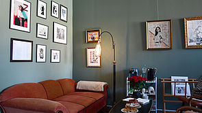 Ein braunes Sof in einer Zimmerecke. Darüber hängen Bilder von Juli Schupa.