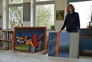 Eine Frau hält ein Bild von Matthias Kanter vor sich. Daneben steht ein weites großes Gemälde. 