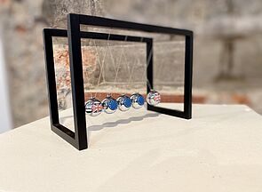 An einem perpetuum mobile hängen fünf silberne Kugeln. Zwei von ihnen tragen die britische Flagge, drei von ihnen die EU-Flagge.