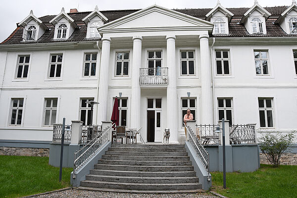 Ein weißes Herrenhaus. Eine große Treppe führt zum Eingang. Dort steht Till Richter mit seinem Hund.