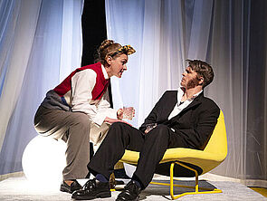 Eine Bühne. Eine Frau sitzt auf einer leuchtenden Kugel. Ihr gegenüber sitzt ein Mann in einem gelben Sessel.