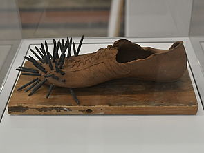 Auf einer Holzplatte steht ein Schuh. Im vorderen Teil des Schuhs stecken Nägel.
