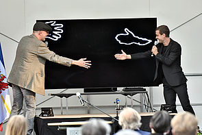 Dr. Marcus Doering und Lars Scheibner stehen auf einer Bühne. Zwischen ihnen steht ein schwarzer Bildschirm. Sie strecken sich die Hände zu, so, als wollten sie sich begrüßen, berühren sich dabei aber nicht. Stattdessen erscheinen auf dem Bildschirm von links und rechts zwei weiße Hände.