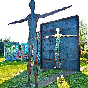 Eine Skulptur in Form eines Menschen. Sie steht mit ausgebreiteten Armen auf einer Wiese. Dahinter steht ein Rechteck, aus dem sie ausgeschnitten zu sein scheint.