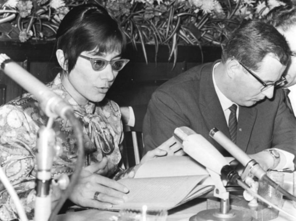 Brigitte Reimann sitzt vor einem aufgeschlagenen Buch und liest. Vor ihr stehen Mikrofone auf dem Tisch. Neben ihr sitzt ein Mann. Das Foto ist eine Schwarz-Weiß-Aufnahme.