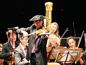 Ein Mann trägt einen Dreieckshut und spielt Geige. Im Hintergrund: die Staatskapelle.