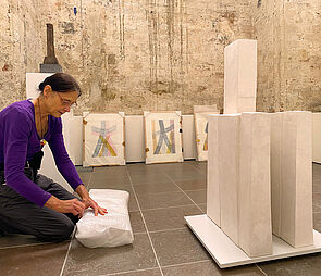 Anne Sewcz kniet vor einer halbfertigen Skulptur aus weißen, rechteckigen und hochkant gestellten Blöcken und öffnet ein Paket, in dem sich weitere Teile der Skulptur befinden. Im Hintergrund lehnen unausgepackte Bilder an der Wand.