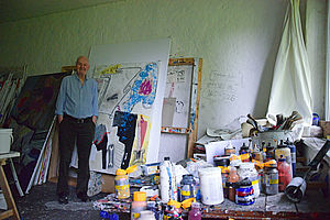 Oscar Manigk steht neben einem mannshohen Gemälde. Auf einem Tisch stehen Farbflaschen und Farbtöpfe.