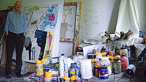 Oscar Manigk steht neben einem mannshohen Gemälde. Auf einem Tisch stehen Farbflaschen und Farbtöpfe.