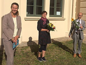 Links steht Ministerin Bettina Martin, in der Mitte, mit Blumen in den Händen, Marion Schael, rechts daneben Dr. Michael Körner. Auch er hält Blumen in der Hand.