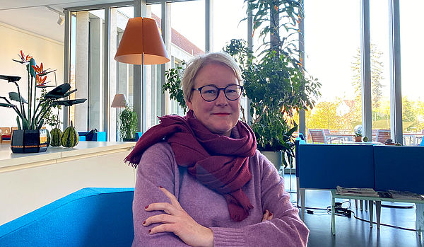 Dorothea Klein-Onnen sitzt auf einem blauen Sofa im Café. im Hintergrund stehen ein Tresen, Tische, Stühle und Pflanzen. Eine raumhohe Fensterwand erhält den Raum.  