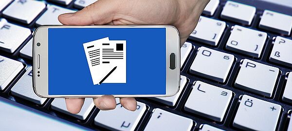 Eine Hand. Die Hand hält ein Handy. Auf dem Handy ist ein Blatt Papier. Auf dem Papier ist Schrift. Hinter der Hand ist eine Tastatur von einem Computer.