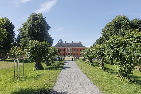 Ein Schotterweg. Links und rechts Wiese mit Bäumen. Am Ende des Weges steht das Schloss Bothmer.