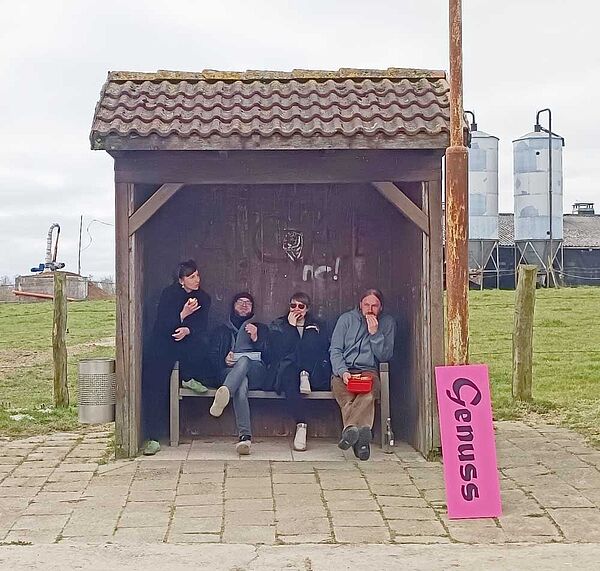 Vier Personen sitzen in einer Haltestelle aus Holz und essen. Neben ihnen lehnt ein Schild mit der Aufschrift "Genuss".