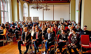 Ungefähr 60 Frauen und Männer sitzen in einem Saal.